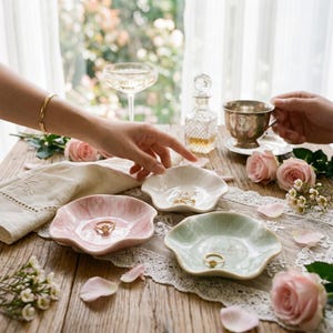 May include: Three small, scalloped-edge ceramic ring dishes in soft pink, white, and pale green. Each dish features gold-colored lettering and holds a gold ring. The dishes are arranged on a wooden surface with lace fabric and pink roses.