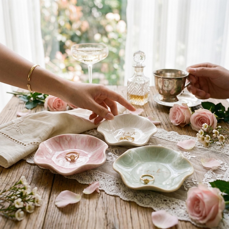 May include: Three small, scalloped-edge ceramic ring dishes in soft pink, white, and pale green. Each dish features gold-colored lettering and holds a gold ring. The dishes are arranged on a wooden surface with lace fabric and pink roses.