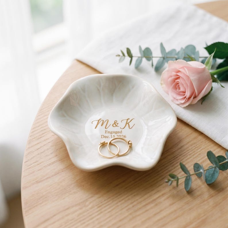 May include: A white ceramic ring dish with a scalloped edge, featuring the gold printed text M & K, Engaged, and Dec. 15 2026. Two gold rings rest inside the dish, which is decorated with a subtle embossed leaf pattern on its interior surface.