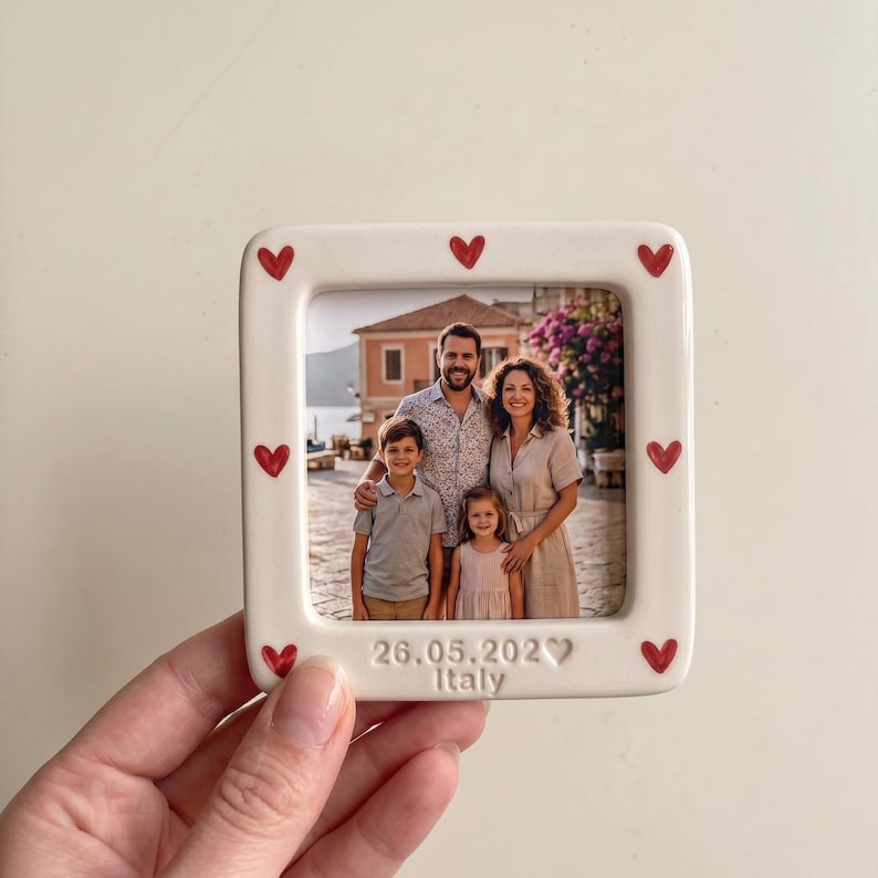 Pode incluir: Uma moldura quadrada de cer&acirc;mica com uma borda cor de creme decorada com pequenos cora&ccedil;&otilde;es vermelhos. A moldura cont&eacute;m uma fotografia de uma fam&iacute;lia de quatro pessoas ao ar livre. A parte inferior tem gravado o texto 26.05.202 e Italy.
