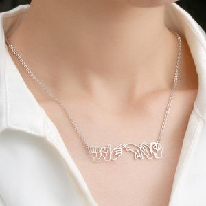Puede incluir: Un collar plateado que muestra la palabra "Love" en lenguaje de señas. El colgante, un diseño calado delicado, cuelga de una fina cadena. El collar se lleva sobre una camisa blanca, creando un contraste.