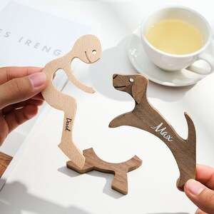 Könnte beinhalten: Holzfiguren einer Person und eines Hundes, mit den Namen "David" und "Max" darauf geschrieben. Die Person ist hell, während der Hund dunkelbraun ist. Ein kleiner Holzsockel ist ebenfalls vorhanden. Eine Tasse Tee ist im Hintergrund.