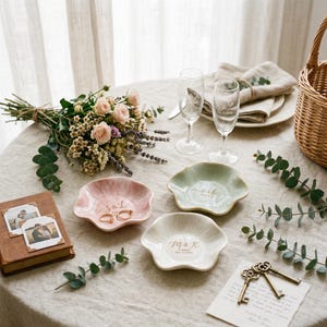 May include: Three scalloped ceramic ring dishes in pastel pink, sage green, and white, each featuring gold-printed initials and dates. The dishes are arranged on a linen-covered table alongside a bouquet of roses, two champagne flutes, and keys.