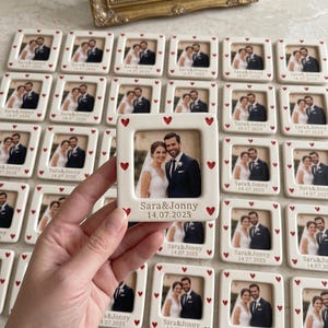 Pode incluir: Uma moldura quadrada de cer&acirc;mica branca com uma fotografia central de um casal em trajes de casamento. A moldura &eacute; decorada com pequenos cora&ccedil;&otilde;es vermelhos ao longo da borda e gravada com o texto "Sara&Jonny" e a data "14.07.2025" abaixo.