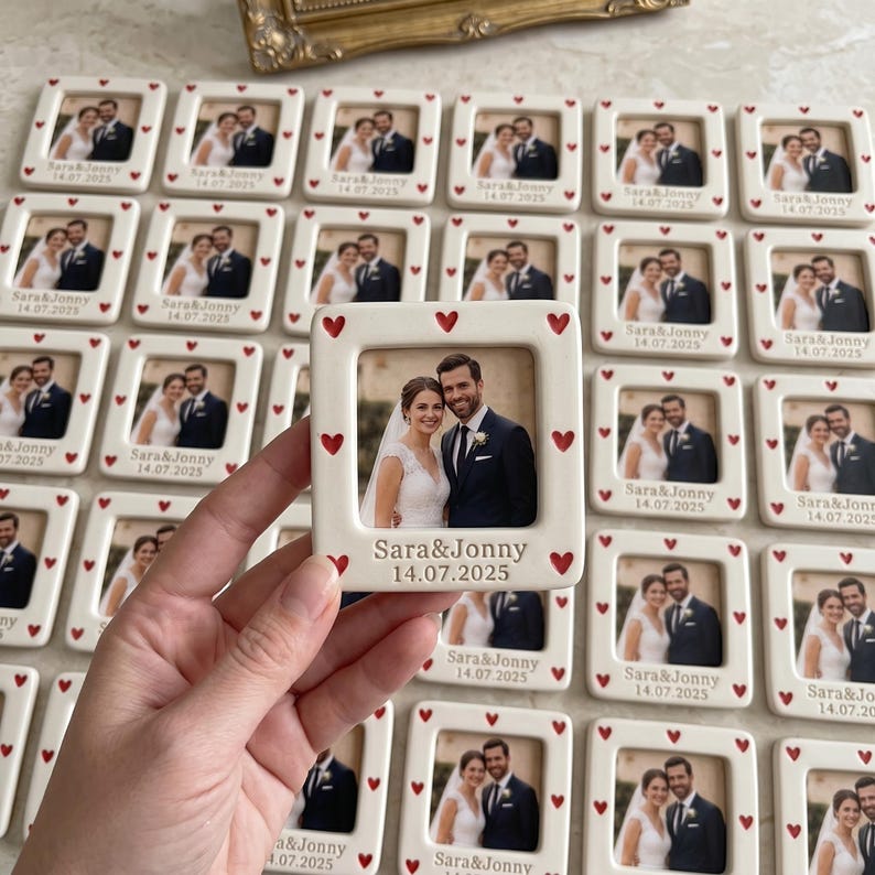 Pode incluir: Uma moldura quadrada de cer&acirc;mica branca com uma fotografia central de um casal em trajes de casamento. A moldura &eacute; decorada com pequenos cora&ccedil;&otilde;es vermelhos ao longo da borda e gravada com o texto "Sara&Jonny" e a data "14.07.2025" abaixo.