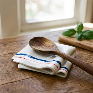 Op de afbeelding: Een houten lepel ligt op een witte keramische lepelhouder met rode en blauwe strepen. De lepelhouder is gevouwen en heeft een gebogen vorm. De lepel is bruin en heeft een lange steel.