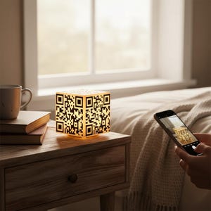 Pode incluir: Luminária em forma de cubo brilhante com design de código QR. A luminária está em uma mesa de cabeceira de madeira ao lado de uma pilha de livros e uma caneca. Um smartphone é segurado, capturando a imagem da luminária.