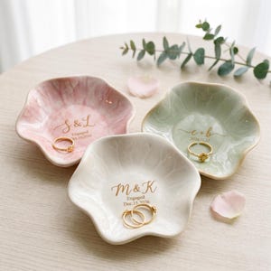 May include: Three scalloped ceramic ring dishes in pastel pink, sage green, and white. Each dish features gold-toned lettering with initials and dates. The pink dish reads S & L Engaged Oct. 10, 2026, the green reads e + b 2026.11.12, and the white reads M & K Engaged Dec. 15, 2026.