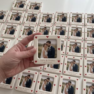 Pode incluir: Uma moldura quadrada de cer&acirc;mica com acabamento branco e pequenos cora&ccedil;&otilde;es vermelhos ao longo da borda. A moldura cont&eacute;m uma fotografia de um casal e tem gravado o texto "Sara & Jonny" e a data "14.07.2025" na parte inferior.