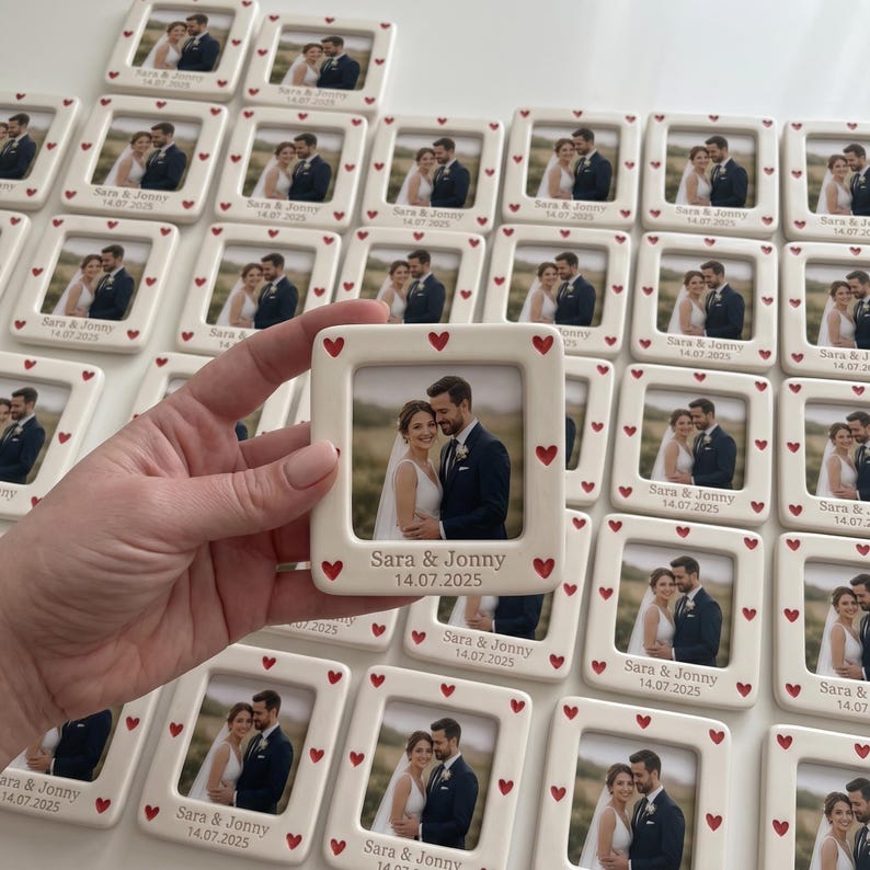 Pode incluir: Uma moldura quadrada de cer&acirc;mica com acabamento branco e pequenos cora&ccedil;&otilde;es vermelhos ao longo da borda. A moldura cont&eacute;m uma fotografia de um casal e tem gravado o texto "Sara & Jonny" e a data "14.07.2025" na parte inferior.
