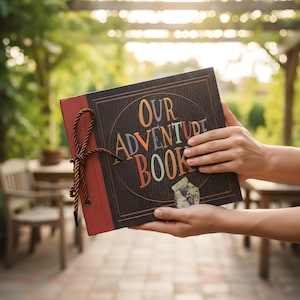 May include: A scrapbook with the words "OUR ADVENTURE BOOK" in colourful letters. The cover is dark brown with a red spine and a brown rope tie. The book is held open, displaying the title.