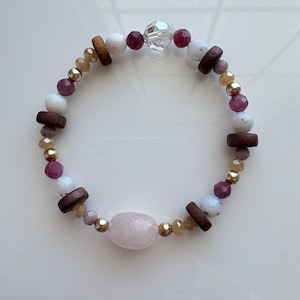 May include: A beaded bracelet with a large, oval pink stone, white, burgundy, and brown beads. Small gold-coloured beads and a faceted clear bead add sparkle. The bracelet is approximately 17.8 cm in circumference.