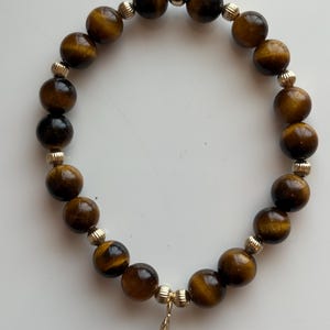 Puede incluir: Una pulsera de cuentas con piedras de ojo de tigre y detalles dorados. La pulsera tiene un dije de hoja y está ensartada en una banda elástica. Las cuentas de ojo de tigre tienen aproximadamente 1 cm de diámetro.