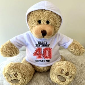 K&ouml;nnte beinhalten: Ein hellbrauner Teddyb&auml;r mit wei&szlig;em Kapuzenpullover und wei&szlig;em T-Shirt. Auf dem T-Shirt steht "HAPPY BIRTHDAY 40 SUZANNE" in Schwarz und Rot. Der B&auml;r hat eine braune Schnauze und Pfoten.