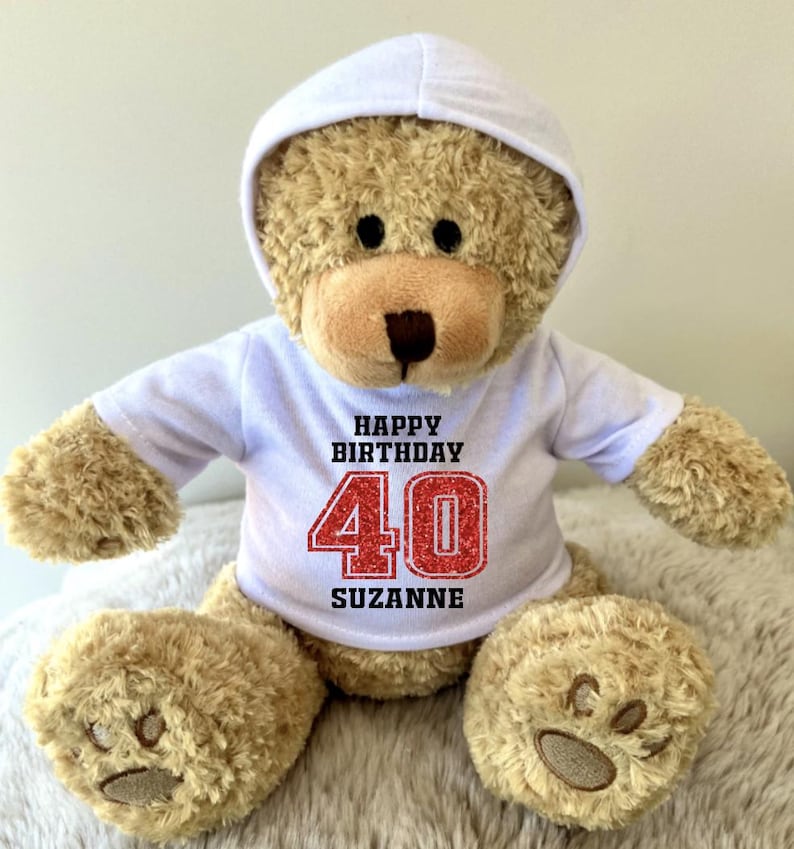 K&ouml;nnte beinhalten: Ein hellbrauner Teddyb&auml;r mit wei&szlig;em Kapuzenpullover und wei&szlig;em T-Shirt. Auf dem T-Shirt steht "HAPPY BIRTHDAY 40 SUZANNE" in Schwarz und Rot. Der B&auml;r hat eine braune Schnauze und Pfoten.