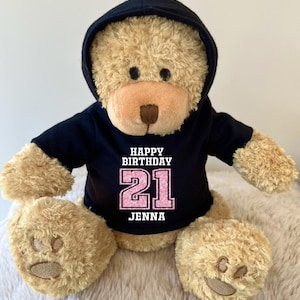 K&ouml;nnte beinhalten: Ein hellbrauner Teddyb&auml;r mit schwarzem Kapuzenpullover. Auf dem Kapuzenpullover steht in Wei&szlig; und Rosa "HAPPY BIRTHDAY 21 JENNA". Der B&auml;r hat eine hellbraune Schnauze und Pfoten.
