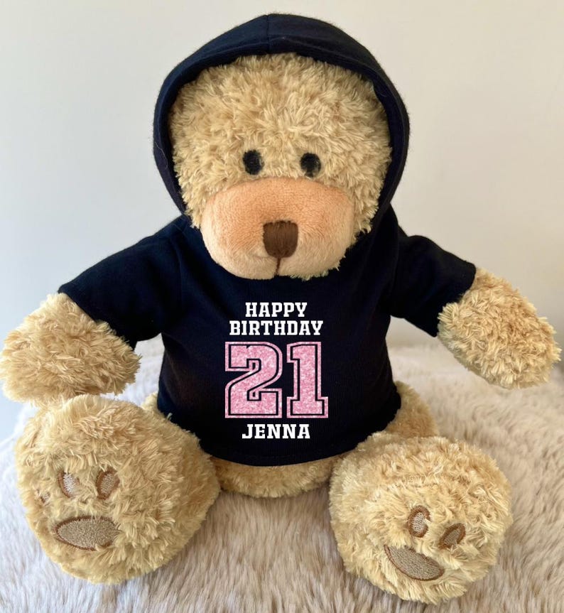 K&ouml;nnte beinhalten: Ein hellbrauner Teddyb&auml;r mit schwarzem Kapuzenpullover. Auf dem Kapuzenpullover steht in Wei&szlig; und Rosa "HAPPY BIRTHDAY 21 JENNA". Der B&auml;r hat eine hellbraune Schnauze und Pfoten.