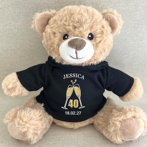 May include: A light brown plush teddy bear wearing a black hooded sweatshirt. The sweatshirt features the name JESSICA, a graphic of two gold glitter champagne flutes, the number 40, and the date 18.02.27 printed in white and gold text.
