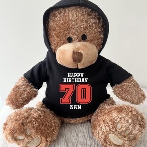 K&ouml;nnte beinhalten: Ein brauner Teddyb&auml;r mit einem schwarzen Kapuzenpullover. Auf dem Kapuzenpullover steht in Wei&szlig; und Rot der Text "HAPPY BIRTHDAY 70 NAN". Der B&auml;r hat eine hellbraune Schnauze und dunkle Augen.