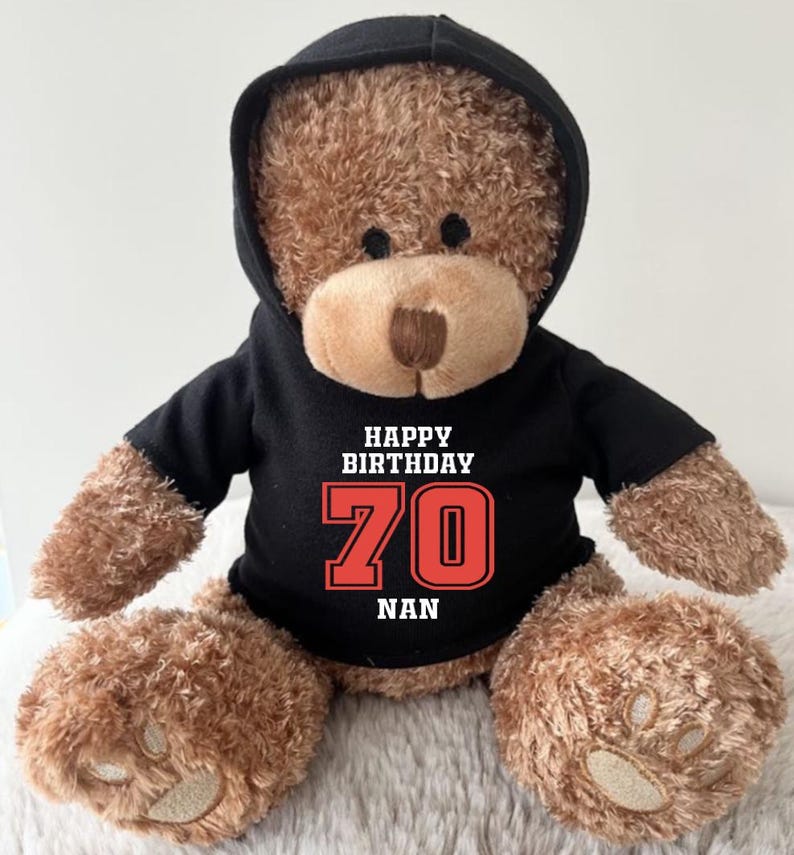 K&ouml;nnte beinhalten: Ein brauner Teddyb&auml;r mit einem schwarzen Kapuzenpullover. Auf dem Kapuzenpullover steht in Wei&szlig; und Rot der Text "HAPPY BIRTHDAY 70 NAN". Der B&auml;r hat eine hellbraune Schnauze und dunkle Augen.