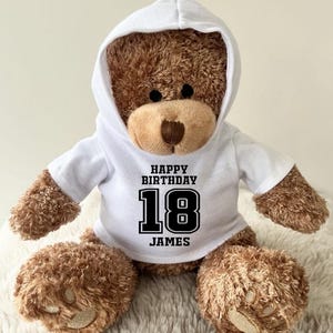 K&ouml;nnte beinhalten: Ein brauner Teddyb&auml;r mit einem wei&szlig;en Kapuzenpullover. Auf dem Sweatshirt steht in schwarzer Schrift "HAPPY BIRTHDAY 18 JAMES". Der B&auml;r sitzt auf einer flauschigen, wei&szlig;en Oberfl&auml;che.