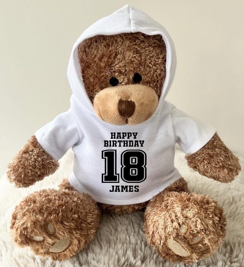 K&ouml;nnte beinhalten: Ein brauner Teddyb&auml;r mit einem wei&szlig;en Kapuzenpullover. Auf dem Sweatshirt steht in schwarzer Schrift "HAPPY BIRTHDAY 18 JAMES". Der B&auml;r sitzt auf einer flauschigen, wei&szlig;en Oberfl&auml;che.