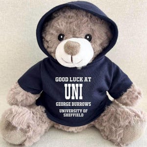 May include: A plush teddy bear with light brown fur wearing a dark navy blue hooded sweatshirt. The hoodie features white text that reads: GOOD LUCK AT UNI, GEORGE BURROWS, UNIVERSITY OF SHEFFIELD. The bear has a white muzzle and dark eyes.