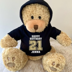 K&ouml;nnte beinhalten: Ein kuscheliger Teddyb&auml;r mit einem schwarzen Kapuzenpullover. Auf dem Pullover steht "HAPPY BIRTHDAY 21 JENNA" in Wei&szlig; und Gold. Der B&auml;r hat hellbraunes Fell und bestickte Pfoten.