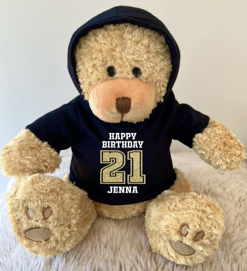 K&ouml;nnte beinhalten: Ein kuscheliger Teddyb&auml;r mit einem schwarzen Kapuzenpullover. Auf dem Pullover steht "HAPPY BIRTHDAY 21 JENNA" in Wei&szlig; und Gold. Der B&auml;r hat hellbraunes Fell und bestickte Pfoten.