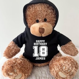K&ouml;nnte beinhalten: Ein brauner Teddyb&auml;r mit einem schwarzen Kapuzenpullover. Auf dem Kapuzenpullover steht in wei&szlig;en Buchstaben "HAPPY BIRTHDAY 18 JAMES". Der B&auml;r sitzt auf einer weichen, wei&szlig;en Oberfl&auml;che.