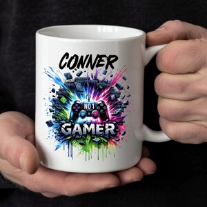 May include: White ceramic mug with the name "CONNER" in black lettering. Below is a colourful graphic of a games controller and the word "GAMER". The design incorporates splashes of blue, green, pink, and purple.