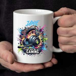 May include: White ceramic mug with a gaming headset design on a brick wall, the name "Jamie", and the text "I can't hear you I'm gaming". The mug has a large handle. The design includes splashes of color.
