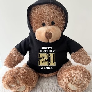 K&ouml;nnte beinhalten: Ein brauner Teddyb&auml;r mit einem schwarzen Kapuzenpullover mit dem Text "HAPPY BIRTHAY 21 JENNA" in Wei&szlig; und Gold. Der B&auml;r hat eine hellbraune Schnauze und sitzt auf einer weichen, wei&szlig;en Oberfl&auml;che.