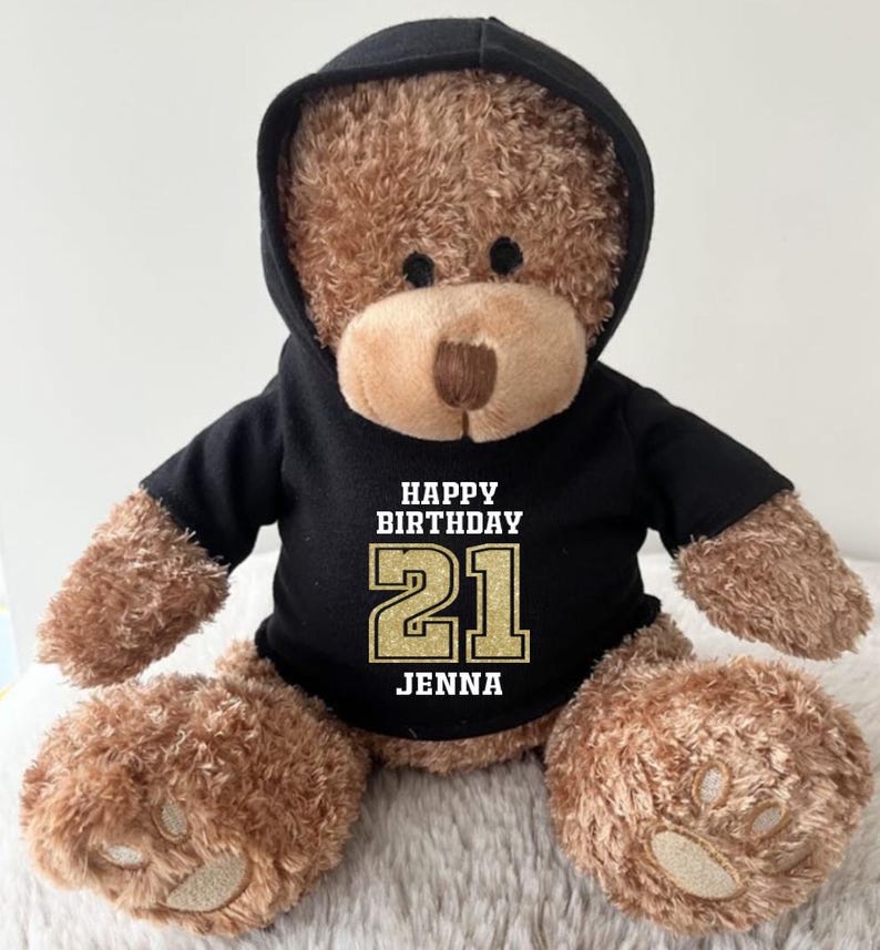K&ouml;nnte beinhalten: Ein brauner Teddyb&auml;r mit einem schwarzen Kapuzenpullover mit dem Text "HAPPY BIRTHAY 21 JENNA" in Wei&szlig; und Gold. Der B&auml;r hat eine hellbraune Schnauze und sitzt auf einer weichen, wei&szlig;en Oberfl&auml;che.