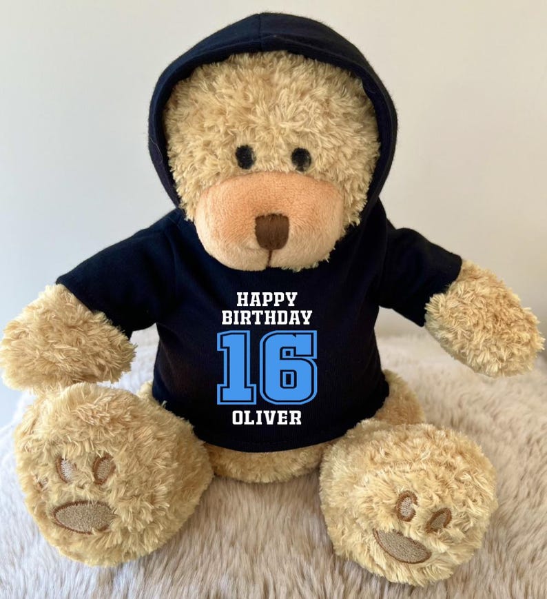 K&ouml;nnte beinhalten: Ein Teddyb&auml;r mit einem schwarzen Kapuzenpullover. Auf dem Kapuzenpullover steht in Wei&szlig; und Blau der Text "HAPPY BIRTHDAY 16 OLIVER". Der B&auml;r hat hellbraunes Fell und gestickte Pfoten.