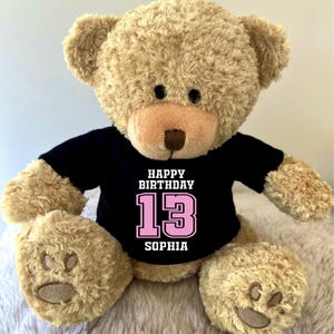 K&ouml;nnte beinhalten: Ein beigefarbener Teddyb&auml;r, der ein schwarzes T-Shirt mit dem Text "HAPPY BIRTHDAY 13 SOPHIA" in Wei&szlig; und Rosa tr&auml;gt. Der B&auml;r hat eine hellbraune Nase und schwarze Augen.