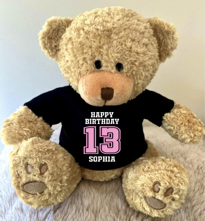 K&ouml;nnte beinhalten: Ein beigefarbener Teddyb&auml;r, der ein schwarzes T-Shirt mit dem Text "HAPPY BIRTHDAY 13 SOPHIA" in Wei&szlig; und Rosa tr&auml;gt. Der B&auml;r hat eine hellbraune Nase und schwarze Augen.