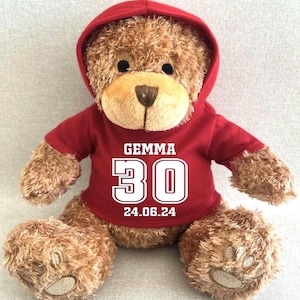 May include: A brown teddy bear wearing a red hoodie. The hoodie displays the name "GEMMA", the number "30", and the date "24.06.24" in white. The bear has a light brown snout and paws, and black eyes. The bear is sitting.
