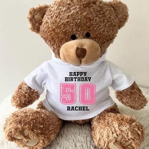 K&ouml;nnte beinhalten: Ein brauner Teddyb&auml;r, der ein wei&szlig;es T-Shirt mit dem Text "HAPPY BIRTHDAY 50 RACHEL" in Schwarz und Pink tr&auml;gt. Der B&auml;r hat eine hellbraune Schnauze und Pfoten und sitzt auf einer flauschigen wei&szlig;en Oberfl&auml;che.