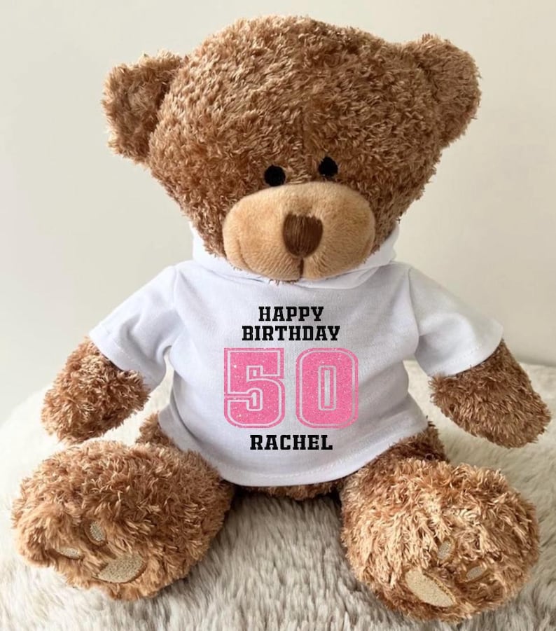 K&ouml;nnte beinhalten: Ein brauner Teddyb&auml;r, der ein wei&szlig;es T-Shirt mit dem Text "HAPPY BIRTHDAY 50 RACHEL" in Schwarz und Pink tr&auml;gt. Der B&auml;r hat eine hellbraune Schnauze und Pfoten und sitzt auf einer flauschigen wei&szlig;en Oberfl&auml;che.