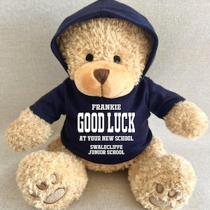May include: A soft teddy bear wearing a navy blue hoodie. The hoodie displays the text "FRANKIE GOOD LUCK AT YOUR NEW SCHOOL SWALCLIFFE JUNIOR SCHOOL" in white. The bear is light brown with a brown nose and black eyes.