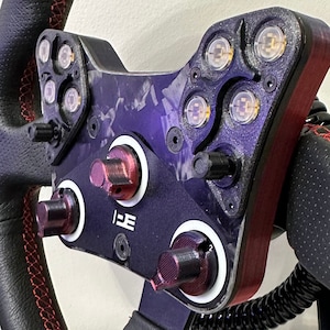 May include: A black leather racing steering wheel with red stitching and a custom-made control panel featuring multiple buttons and lights. The wheel is mounted on a black stand.