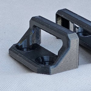 May include: Two black plastic 3D printed brackets with a rectangular opening in the center. The brackets have a beveled edge and two small holes on the bottom.