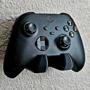 May include: A black Xbox Series X controller with a black wall mount. The controller has a directional pad, four face buttons, two analog sticks, and two triggers.
