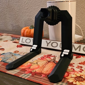 May include: A black 3D printed phone stand with a gear-like design on the top. The stand is on a table with a fall-themed table runner.