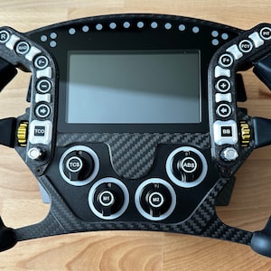 Peut inclure: Un volant de course noir et en fibre de carbone avec plusieurs boutons et un écran central. Le volant possède des poignées ergonomiques et divers boutons de commande avec des abréviations telles que "RAD", "TCS" et "ABS".