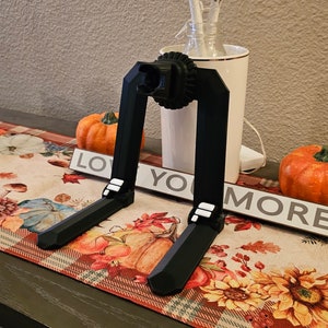 May include: A black 3D printed phone stand with a circular base and two adjustable legs. The stand is on a table with a fall-themed table runner. The table runner has a red and white checkered pattern with fall leaves and pumpkins.