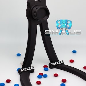 May include: A black 3D printed stand with the word "MOZA" printed on each leg. The stand is designed to hold a racing wheel and has a round opening at the top. The stand is on a white surface with scattered blue and red candies.