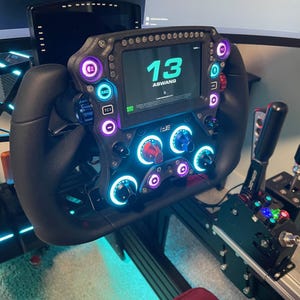 May include: A black racing steering wheel with a digital display showing "13 ASWAND". The wheel has multiple buttons and knobs with colorful LED lights.
