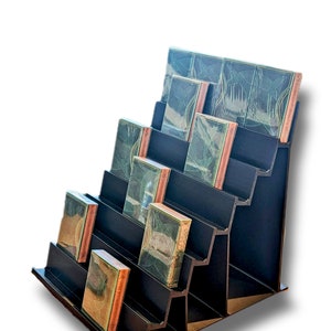 May include: A black display stand with multiple tiers holds a collection of small, rectangular books with green and brown covers. The books are arranged in a staggered pattern on the stand.